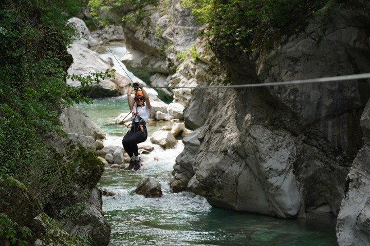 A smiling woman ziplining across a deep, rocky canyon with a turquoise river and lush vegetation in the background.