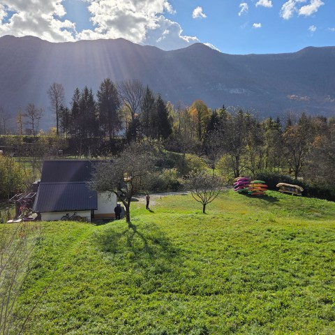 Scenic mountain view with a house, trees, and kayaks on a grassy field under a partly cloudy sky.